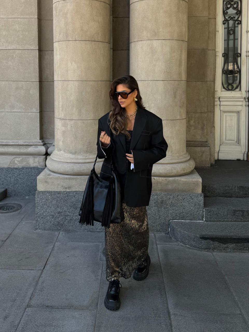 Look com blazer preto e saia longa estampada diante de colunas de pedra
